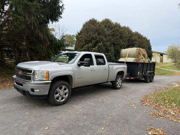 Silver pickup truck towing a trailer loaded with hay bales on a paved driveway.
