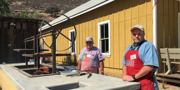 Two men grilling meat outdoors near a yellow building.
