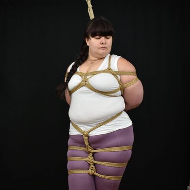 Black background; brunette femme wearing white shirt & pink pants has rope on chest and thighs