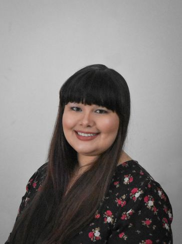 A smiling, White person with long, dark brown hair with bangs and brown eyes, in a floral shirt.