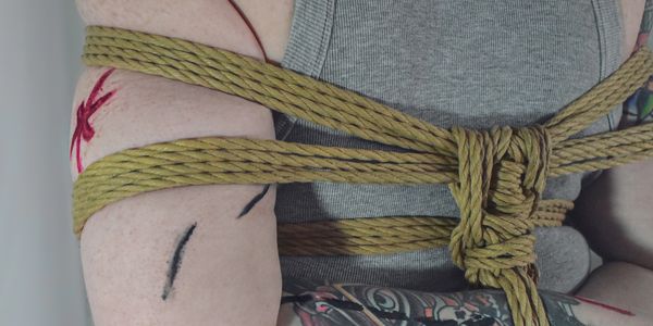 A White person wearing a gray tank top is tied with ropes with their arms behind their back.