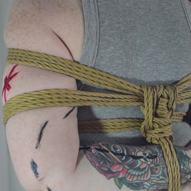 A white person with arms crossed behind their back. Rope is tied across their arms and chest and aro