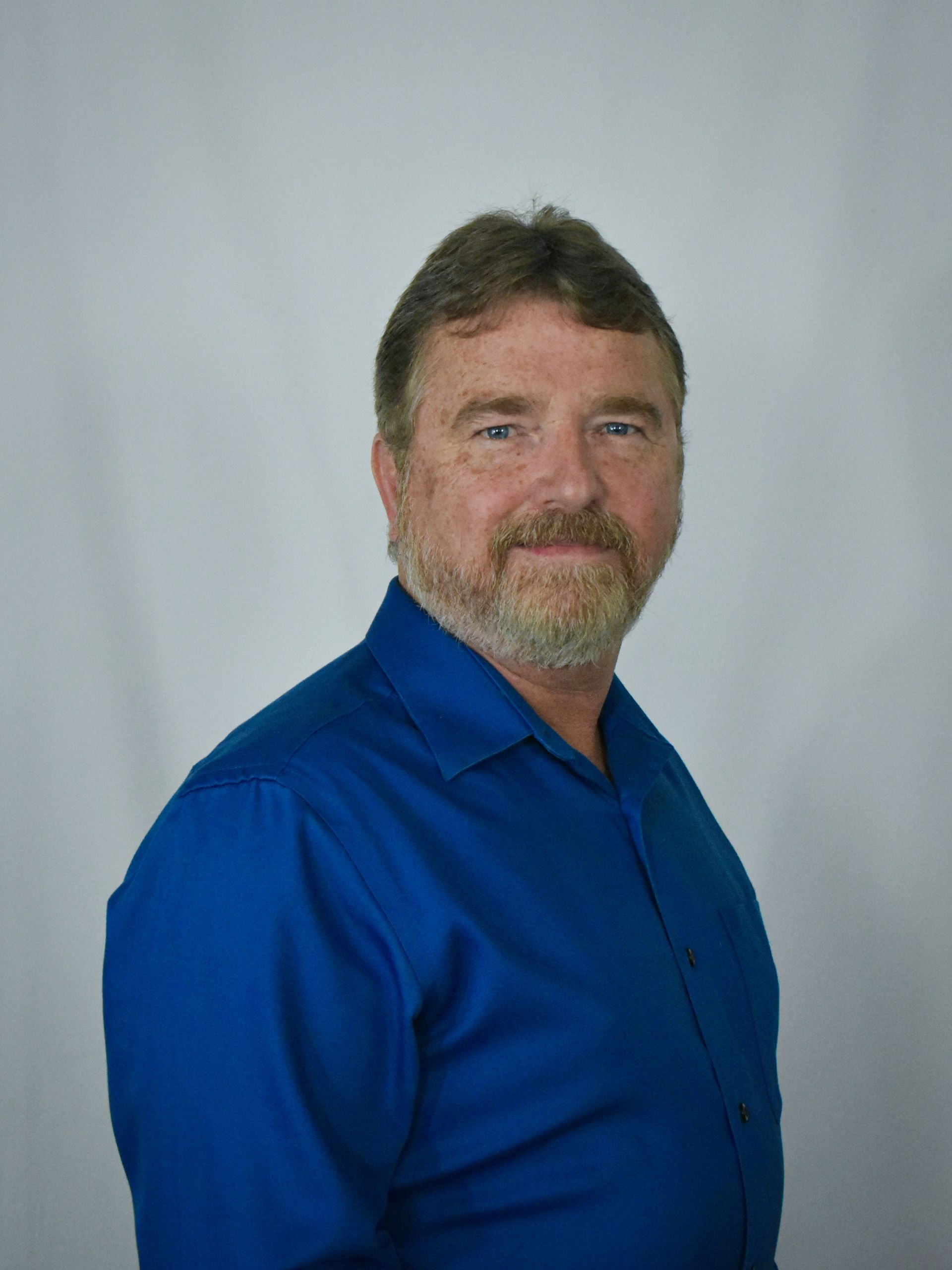 A middle-aged white man with brown hair and a gray beard wearing a blue shirt, white background