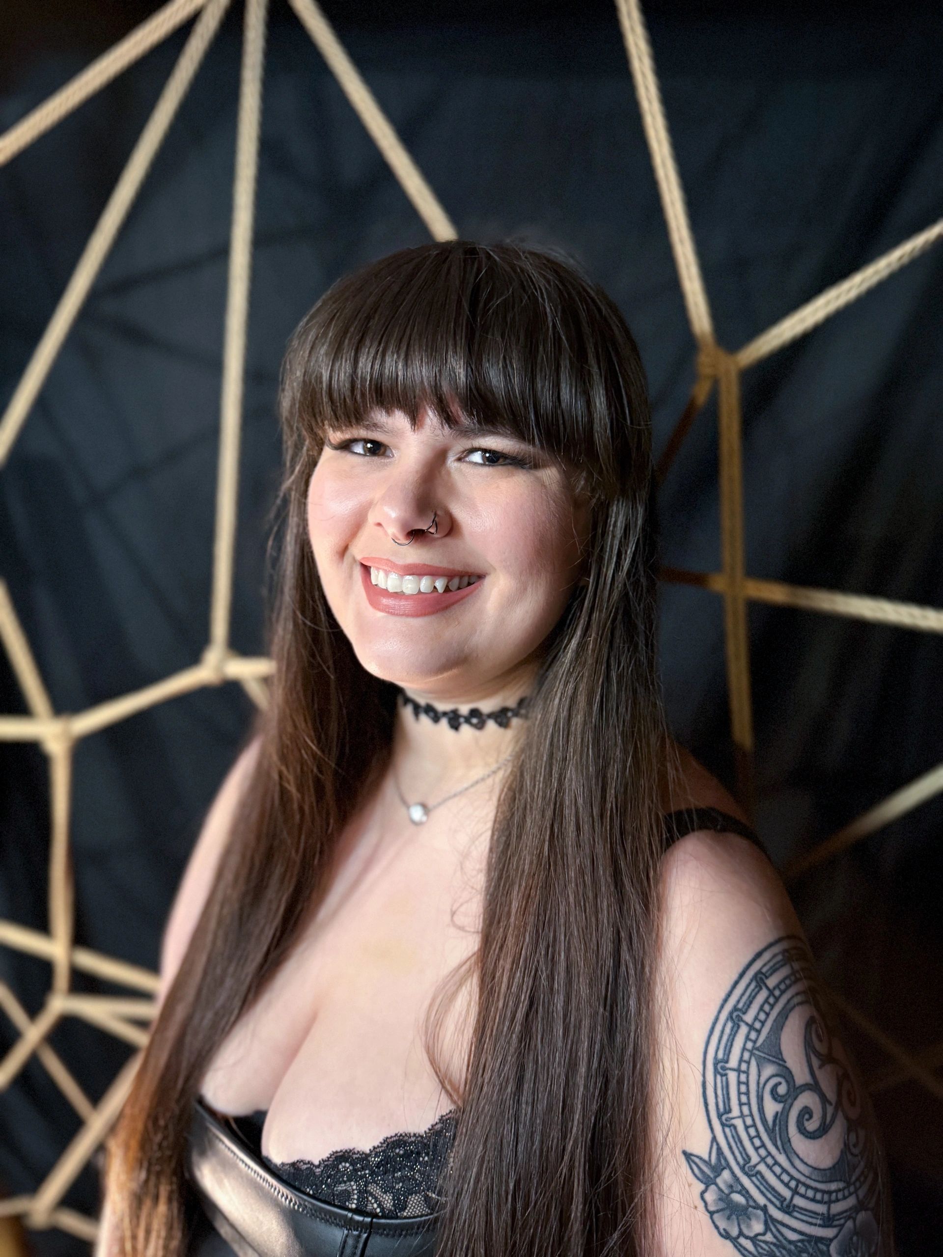 A white non-binary femme with a tattoo, nose piercings, long brown hair, & a black top is smiling.