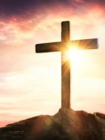 Wooden cross on a hill with sun shining behind it at sunset.