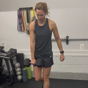 Woman exercising with dumbbell in a gym wearing black workout clothes.