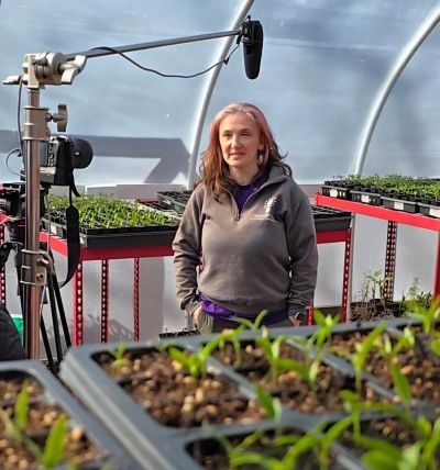 Woman standing in greenhouse surrounded by plants and filming equipment.