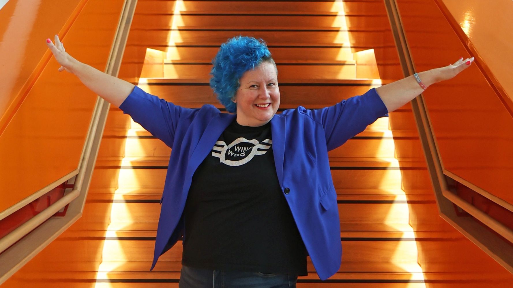 Wing Woman Plus founder, Lynnette Ellison, with a blue blazer, black tee and blue side shave hair