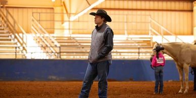 Man in cowboy hat standing in arena with horse and child.