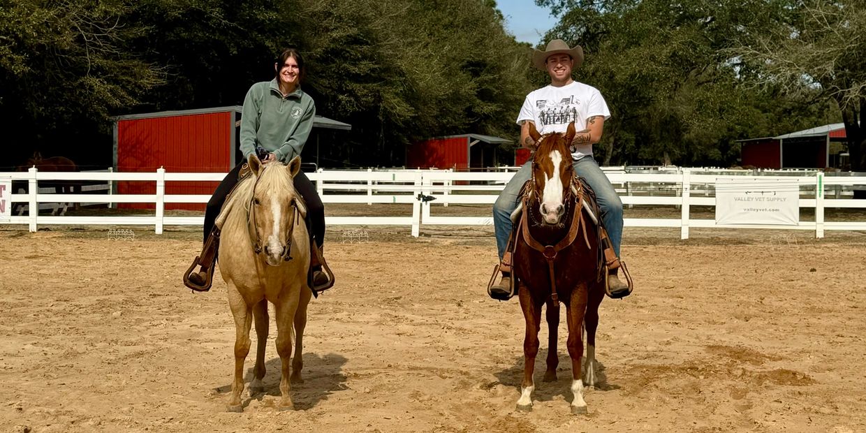 Two people smiling while riding horses in an outdoor arena.