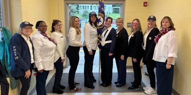 A group of nine women standing indoors posing for a photo, some wearing uniforms and hats.