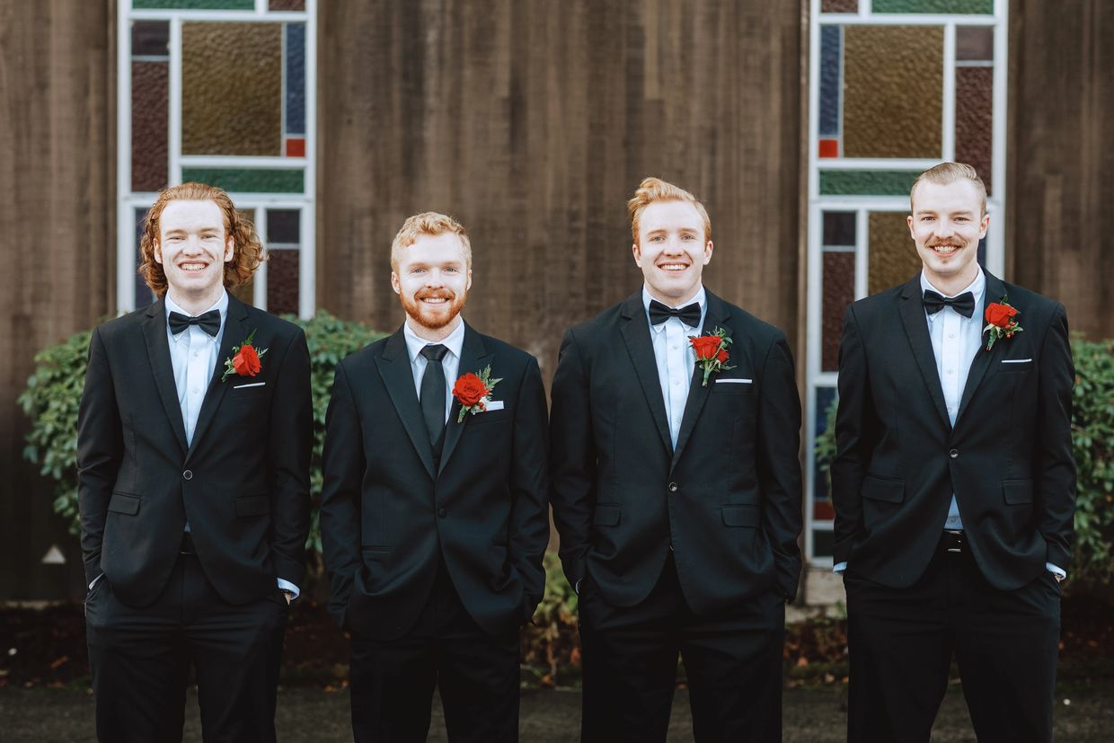 Four men in tuxedos with red boutonnieres smiling outdoors.