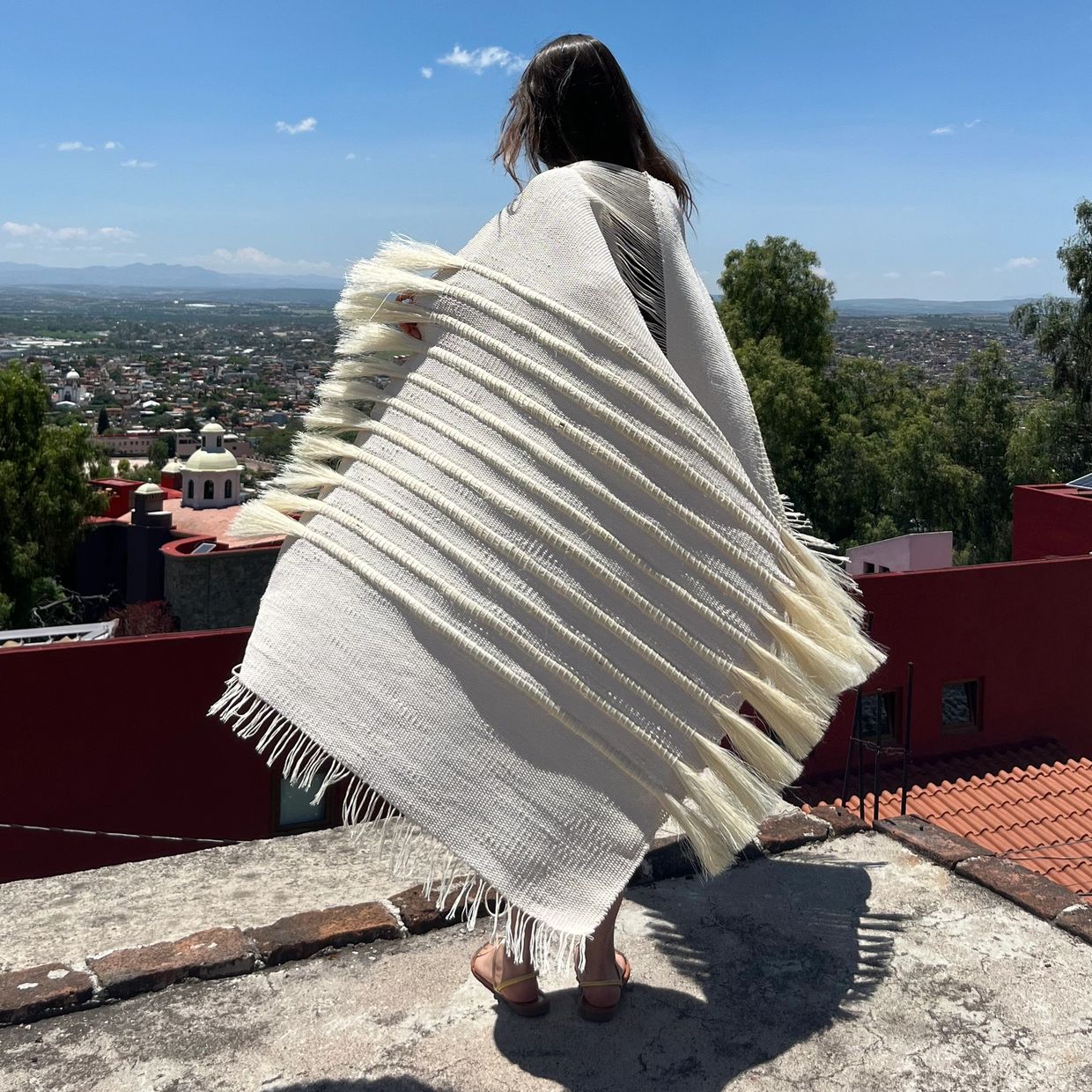 Woman wrapped in a fringed white shawl overlooking a cityscape on a sunny day.