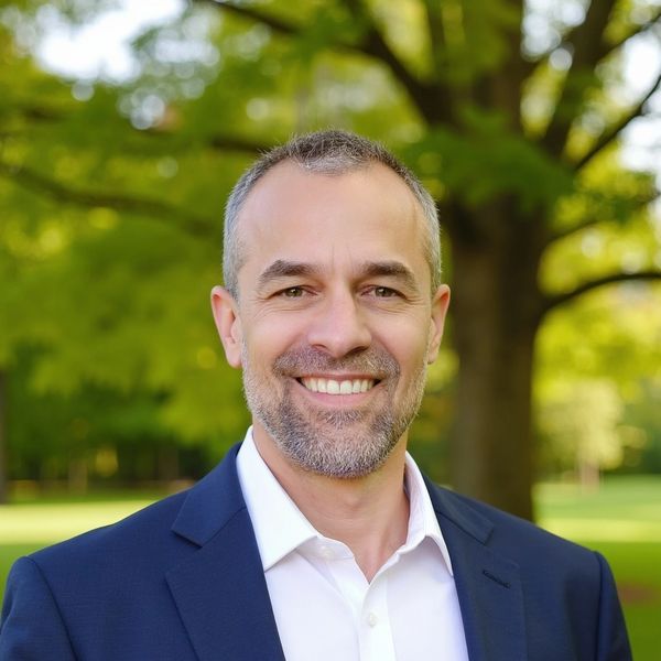 Smiling man in a navy suit outdoors with green trees in the background.