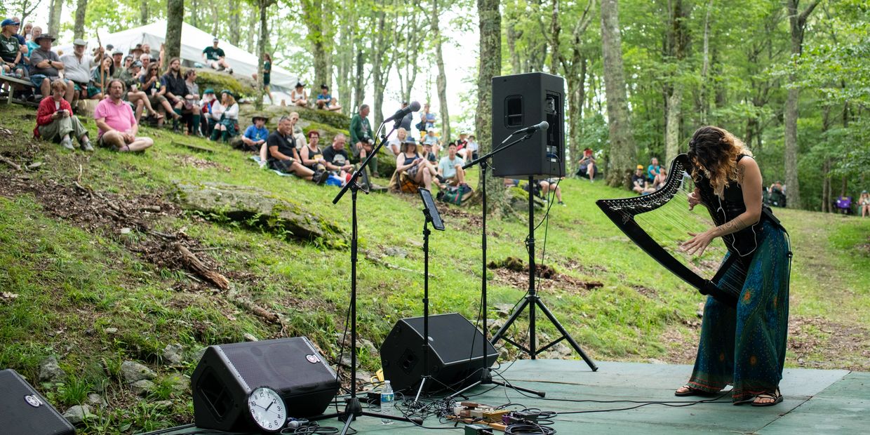 A woman plays a harp on an outdoor stage with an audience seated on a grassy hill.