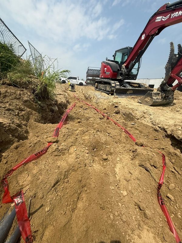 Excavator at a construction site with a trench marked by red warning tape.