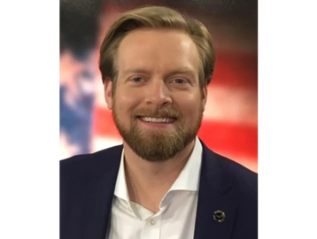 Smiling man with beard in suit against blurred American flag background.
