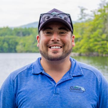 Smiling man in blue polo shirt and cap by a lake.