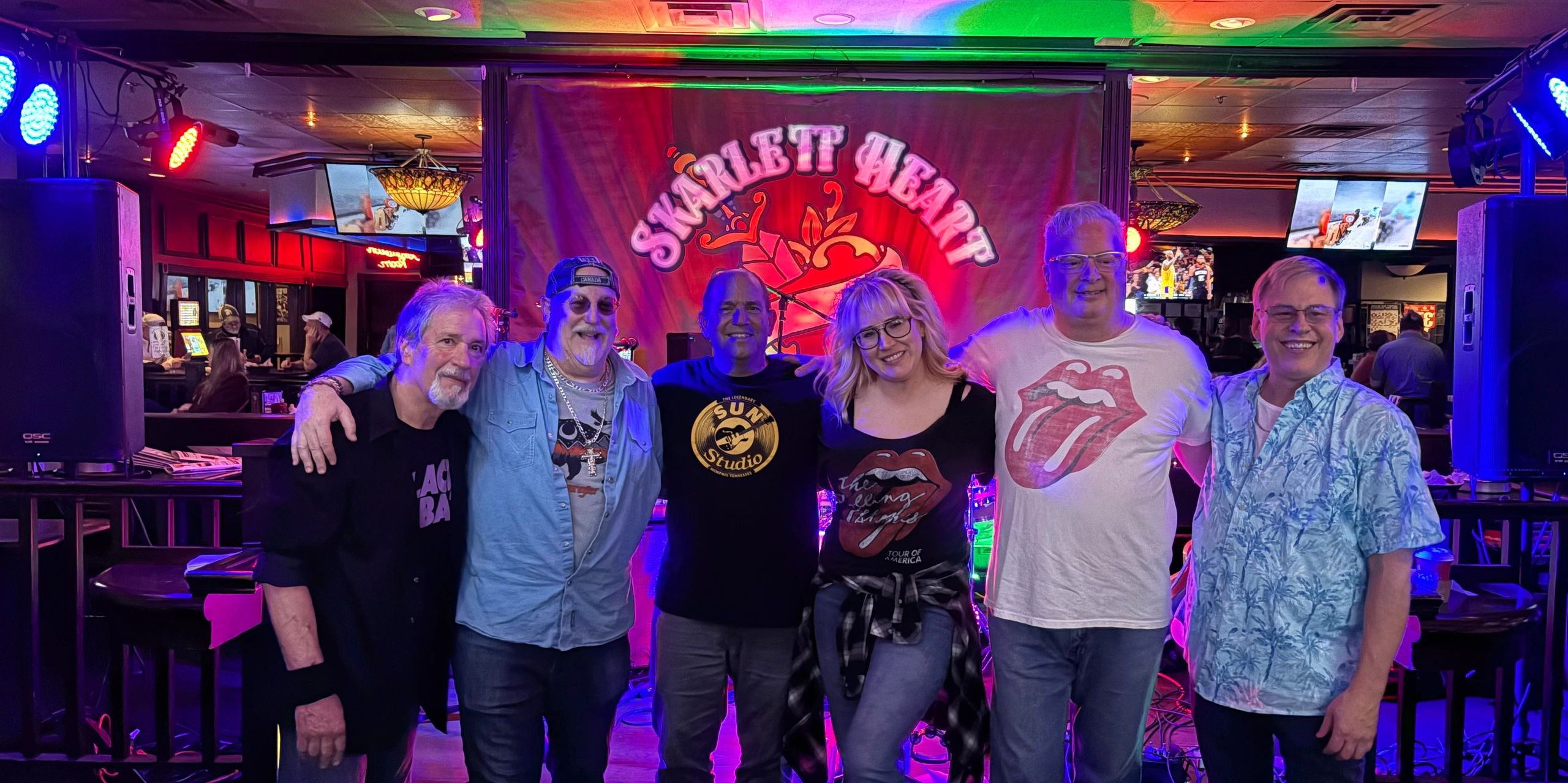 A group of six people smiling in front of a Scarlet Heart banner in a lively bar setting.