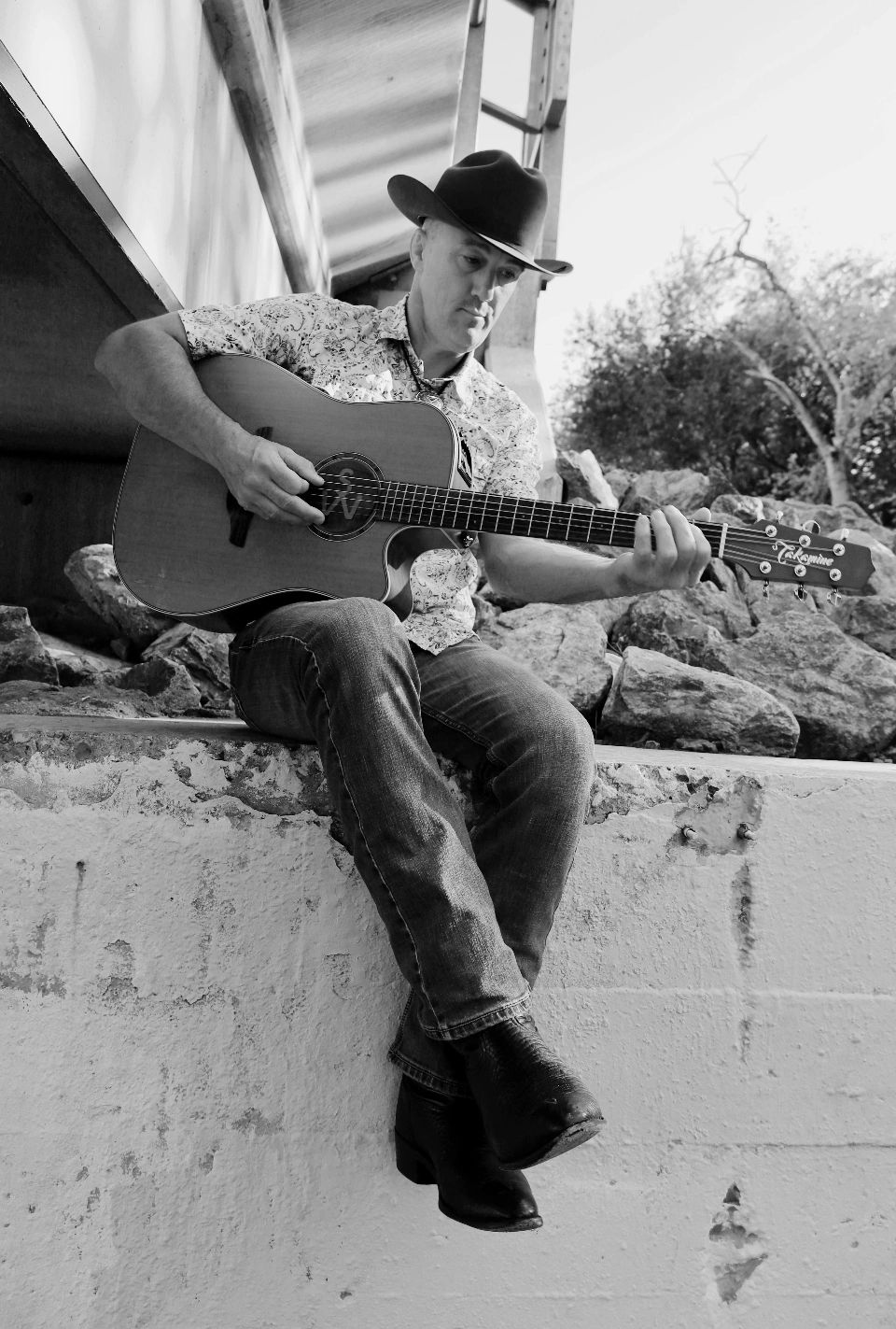 Man in cowboy hat playing guitar outdoors on a concrete ledge.