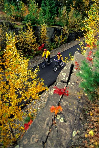 Two cyclists ride on a paved path surrounded by colorful autumn trees.