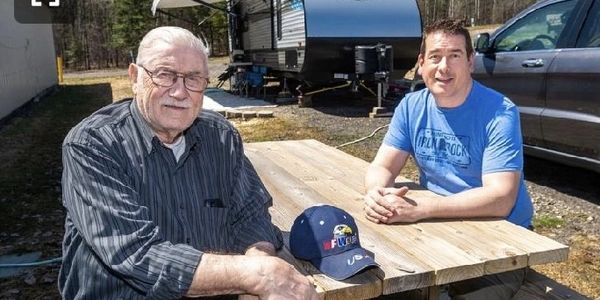 Two men sitting at a wooden picnic table outdoors near a camper trailer.
