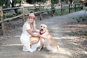 Natural light portrait of a girl in a white dress and tan hat kneeling on a path hugging her dog.