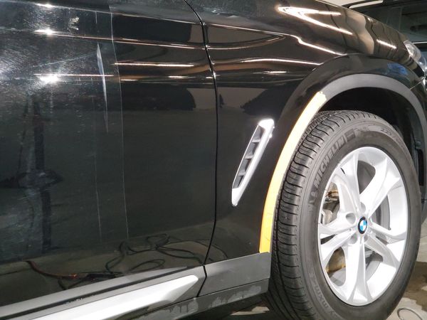 "Close-up of a black car's side panel and wheel, showing light reflections and paint imperfections."