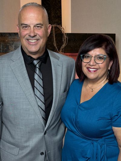 A man in a gray suit and a woman in a blue dress smiling together indoors.