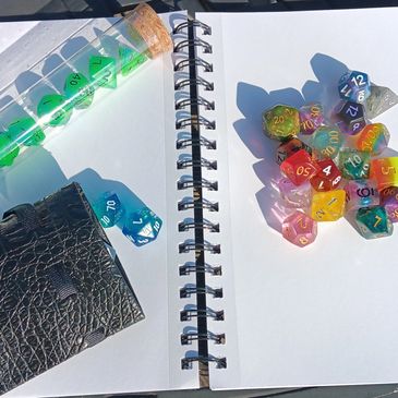 Journal with plain pages opened. On top rests a tube of dice, a dice bag, and a handful of random die