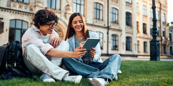 Two students sitting on grass, smiling and using a tablet outside a historic building.
