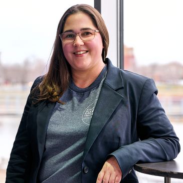 Femme souriante avec lunettes et blazer près d'une fenêtre.