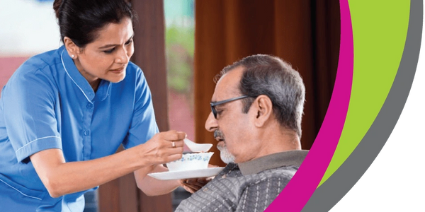 Caregiver in blue uniform feeding an elderly man with a spoon.