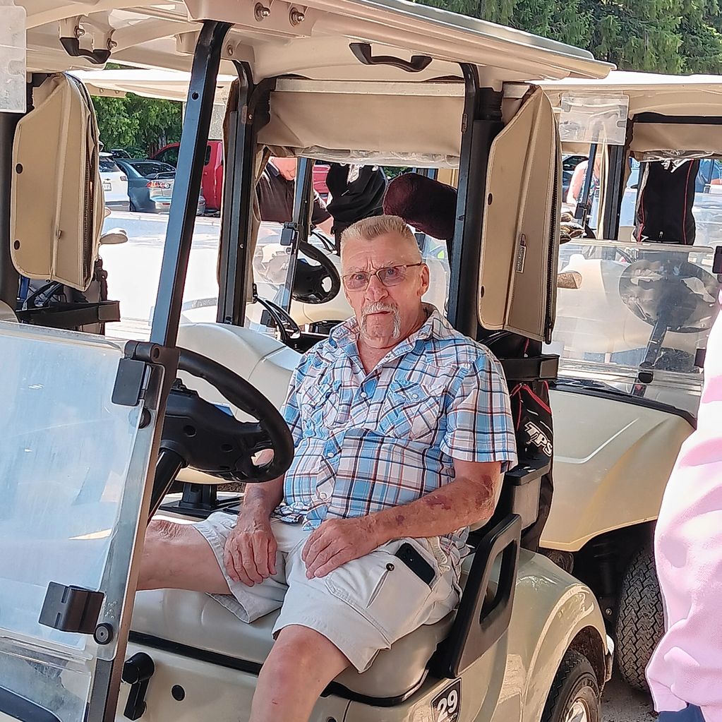 Man sitting in a golf cart wearing glasses and a plaid shirt.