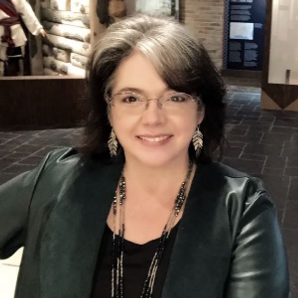 A woman with brown and silver hair wearing large silver earrings, smiles towards the viewer.