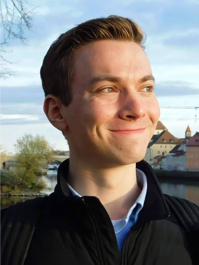 A young man smiling outdoors with a scenic background of buildings and water.