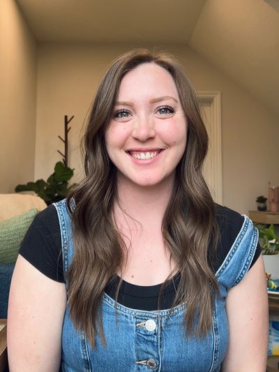 A smiling woman with long brown hair wearing a denim dress over a black shirt.