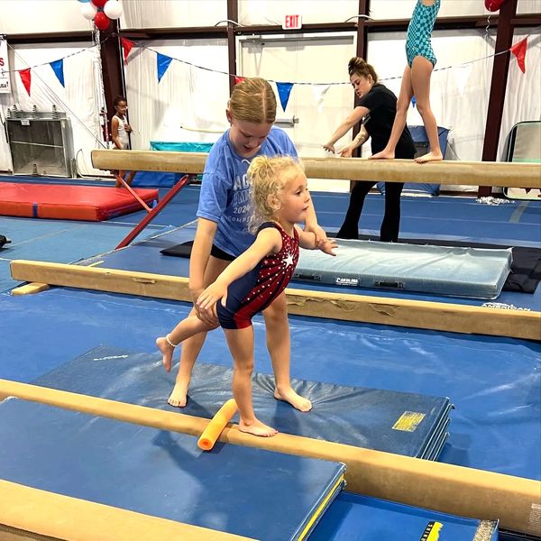 A young girl practices balance on a beam with assistance in a gym.