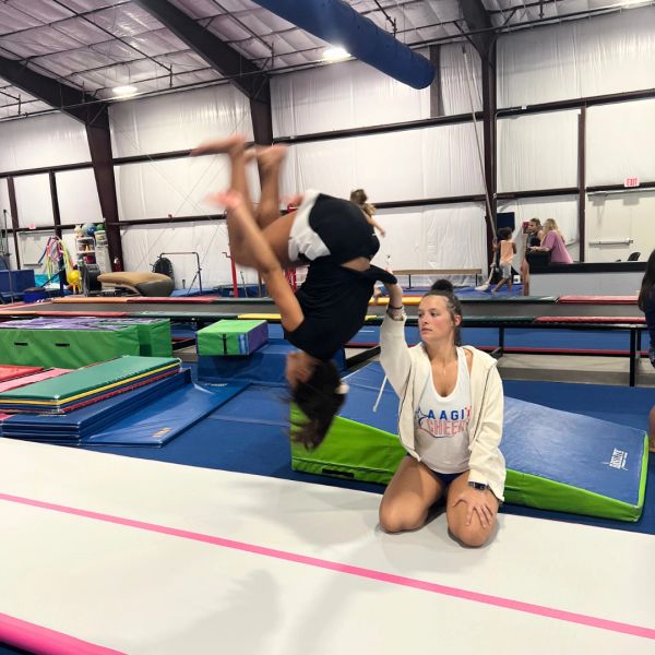 Coach assists gymnast in mid-flip at training gym.