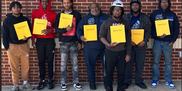 Seven men standing in front of a brick wall holding yellow folders labeled Appalachian.