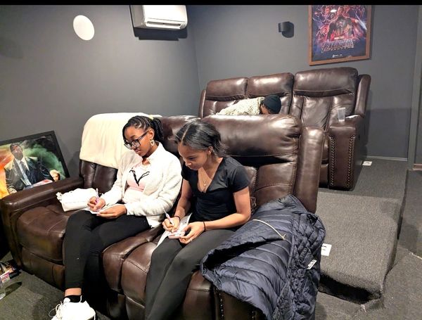 Two girls writing on notepads in a cozy home theater with recliner seats.