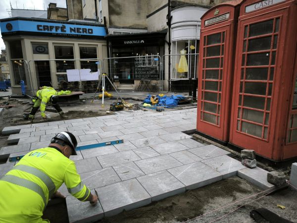 Workers laying pavement near red phone boxes and a cafe.