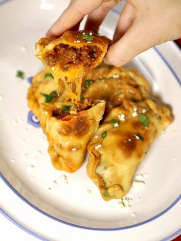 Hand holding a cheesy, meat-filled empanada with more empanadas on a plate.