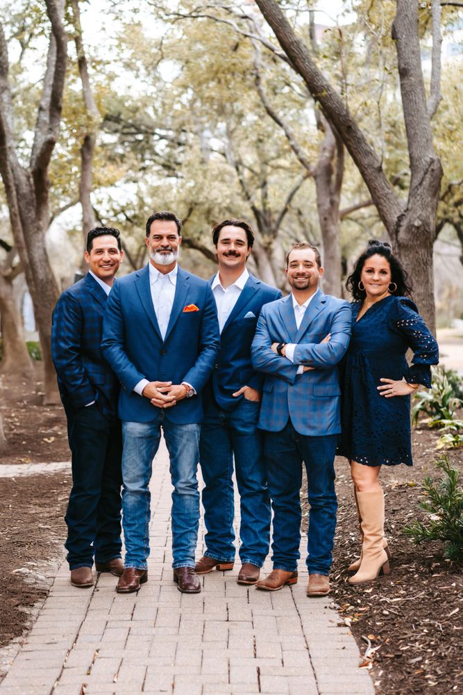 Group of five adults dressed in blue, posing outdoors on a pathway surrounded by trees.