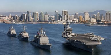 A naval fleet with an aircraft carrier and destroyers near a city skyline.