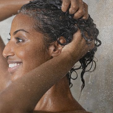 Mulher de pele morena, sorridente lavando os cabelos com shampoo natural, sem ingredientes sintético