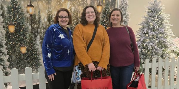 Three women smiling, holding shopping bags in a festive winter setting with decorated Christmas trees.