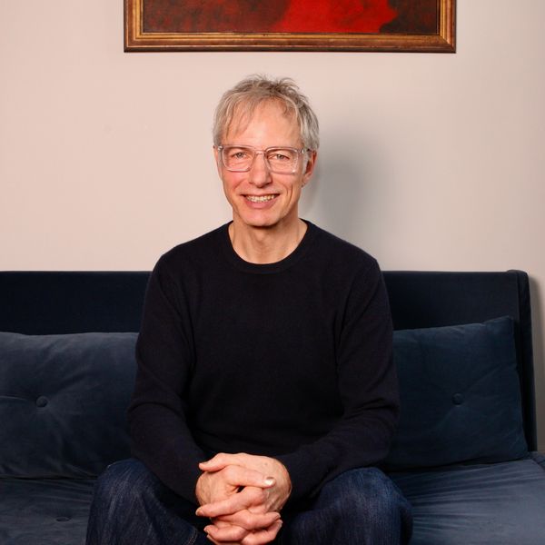 Smiling man with glasses sitting on a dark couch under a red painting.