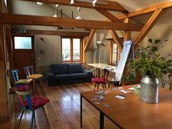 ein Seminarraum mit einem Stuhlkreis, einer Flipchart, einem Blumenstrauß und Karten auf einem Tisch