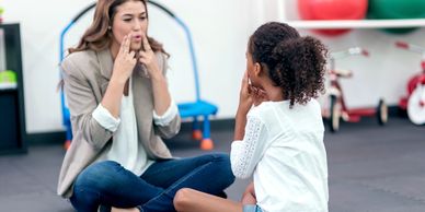 Girl sits on floor facing female Speech Therapist and holds fingers on face while practicing speech